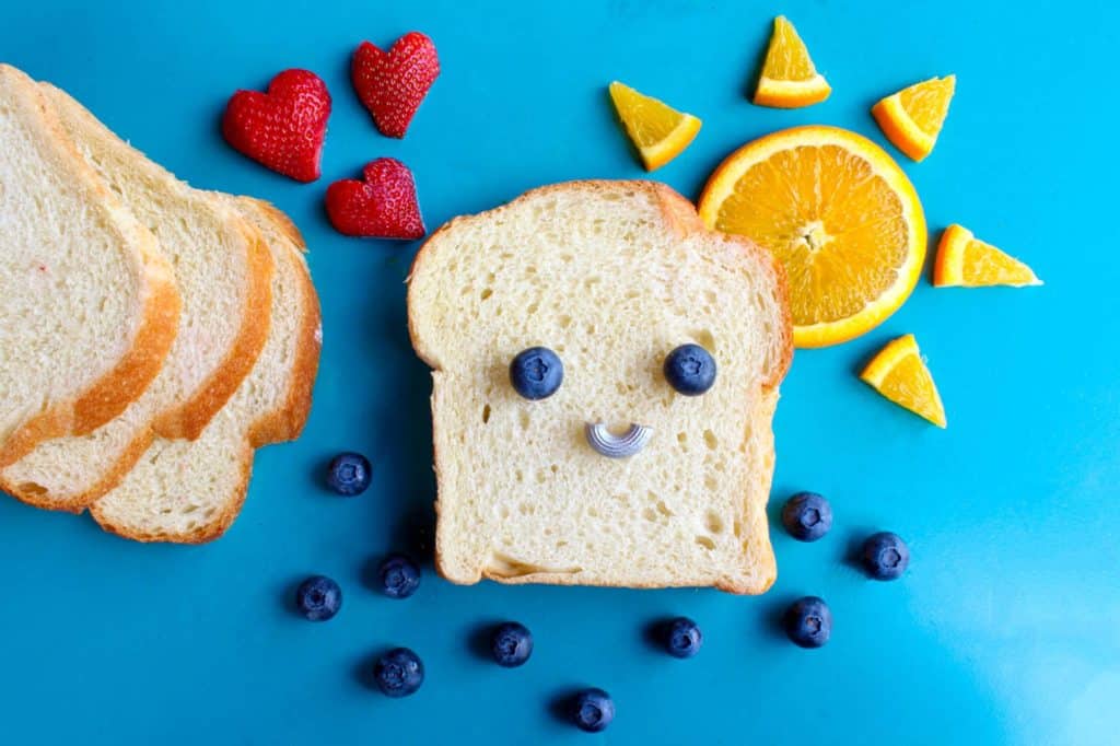 Slice of bread with blueberry eyes with an orange looking like the sun and strawberries in shapes of hearts