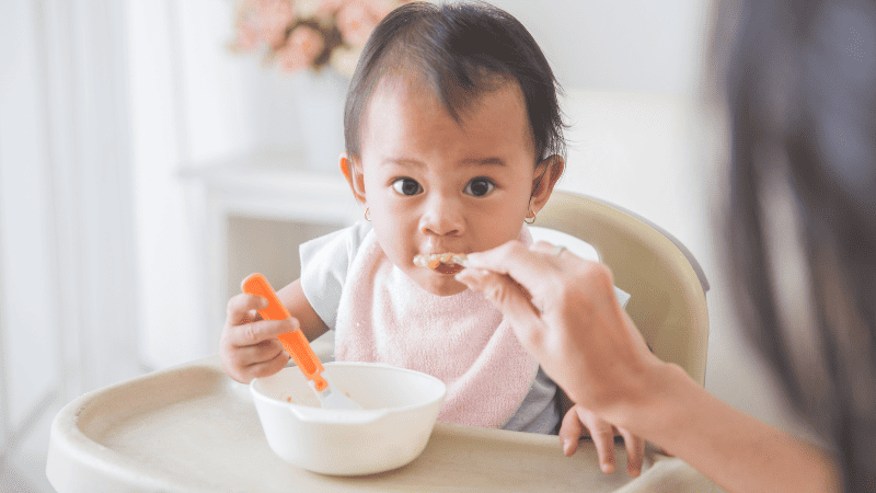 Baby sitting in a highchair eating and holding spoon