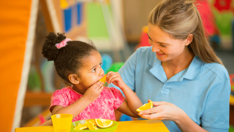 A Nursery Practitioner Sits and Eats Oranges with a Young Child In Nursery School